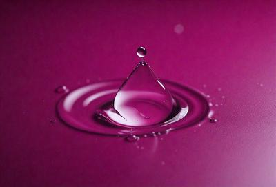 The primary subject of the image is a single drop of water, which is captured in a close-up shot. The setting is a pink surface, which adds a vibrant and warm atmosphere to the scene. The visual style is a realistic representation of the water drop, and the colors and mood are predominantly pink, creating a sense of warmth and tranquility.
