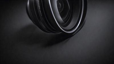 The primary subject of the image is a close-up of a lens, which is the main object in the scene. The lens is placed on a black background, creating a contrast between the lens and the setting. The visual style of the image is a black and white photo, giving it a classic and timeless appearance. The colors and mood of the image are predominantly black, with the lens being the only object with a reflective surface, adding a touch of brightness to the scene.
