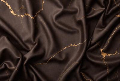 The primary subject of the image is a piece of fabric, possibly a piece of clothing or a curtain, with a brown color and a pattern of gold and brown. The setting is a room with a wall, and the visual style appears to be a close-up of the fabric. The colors and mood of the image are warm and earthy, with a sense of luxury and elegance.