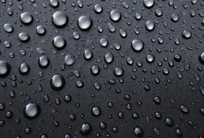 The primary subject of the image is a close-up of a black surface with numerous small droplets of water on it. The setting appears to be a dark room, and the visual style is a close-up of the water droplets. The colors and mood of the image are predominantly dark, with the droplets of water being the main focus.