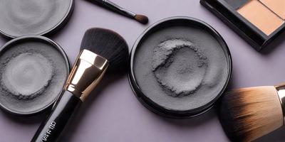 The primary subject of the image is a black jar of makeup, which is placed on a table. The jar is surrounded by various makeup tools, including a brush and a spoon. The setting appears to be a workspace or a makeup station, with a gray background that complements the black jar. The visual style of the image is a close-up of the makeup jar and tools, emphasizing the details of the makeup products and tools. The colors and mood of the image are predominantly dark,