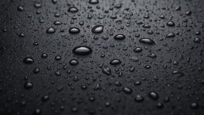 The primary subject of the image is a close-up of a black surface with numerous droplets of water on it. The setting is a dark background, and the visual style appears to be a photo. The colors and mood of the image are predominantly dark, with the droplets of water being the main focus.