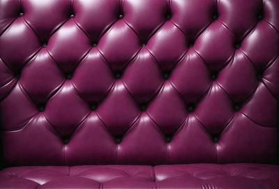 The primary subject of the image is a purple leather couch with a purple velvet cushion. The setting is a room with a purple wall, which complements the couch's color. The visual style is a close-up of the couch, showcasing its design and texture. The colors and mood of the image are predominantly purple, creating a cozy and inviting atmosphere.