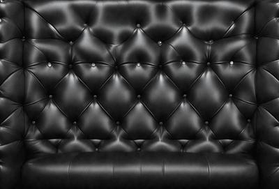 The primary subject of the image is a black leather couch. The setting is a room with a black background, which creates a contrast with the couch. The visual style is a black and white photo, giving the image a classic and timeless appearance. The colors and mood of the image are predominantly black, creating a sense of sophistication and elegance.
