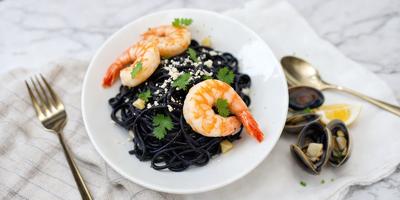 The primary subject of the image is a bowl filled with a delicious meal consisting of shrimp and spaghetti. The bowl is placed on a dining table, and the shrimp and spaghetti are arranged in a visually appealing manner. The setting includes a white plate and a spoon, which are also present on the table. The visual style of the image is a close-up of the bowl, showcasing the details of the meal. The colors and mood of the image
