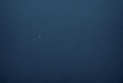 The primary subject of the image is a moon, which is visible in the sky. The setting is a dark, cloudy night with a blue sky. The visual style is a photo, capturing the moon and the night sky. The colors and mood of the image are predominantly dark and moody, with the moon being the main source of light in the scene.