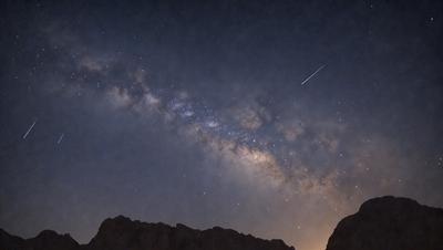 The primary subject of the image is the night sky filled with stars. The setting is a dark, cloudy night with a rocket launching into the sky. The visual style is a photo of the night sky, capturing the beauty of the stars and the rocket's trajectory. The colors and mood of the image are dark and dramatic, with the bright stars and the rocket's light creating a sense of wonder and excitement.