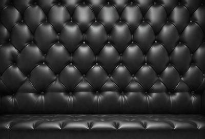 The primary subject of the image is a black leather couch. The setting is a black background, which creates a striking contrast with the couch. The visual style is a close-up of the couch, showcasing its texture and details. The colors and mood of the image are predominantly black, giving it a sleek and modern appearance.