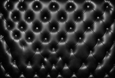 The primary subject of the image is a black leather couch. The setting is a black background, which creates a striking contrast with the couch. The visual style is a close-up of the couch, showcasing its texture and details. The colors and mood of the image are predominantly black, giving it a sleek and modern appearance.
