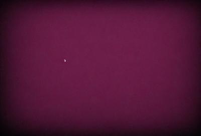 The primary subject of the image is a small, bright moon in the sky. The setting is a dark, purple background, which creates a contrasting and dramatic effect. The visual style is a photo, capturing the moon in its natural environment. The colors and mood of the image are predominantly dark and moody, with the purple background adding a sense of depth and intensity.