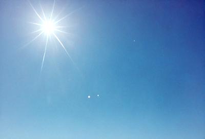 The primary subject of the image is the sun, which is shining brightly in the sky. The setting is a clear blue sky, with no clouds or other obstructions visible. The visual style of the image is a photo, capturing the sun's rays and the vast expanse of the sky. The colors in the image are predominantly blue, with the sun being the main source of light and warmth. The overall atmosphere of the image is serene and peaceful, as the sun shines down on the