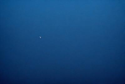 The primary subject of the image is the moon, which is seen in the sky. The setting is a dark blue sky, and the visual style is a photo. The colors and mood of the image are predominantly dark and moody, with the moon being the main source of light in the scene.