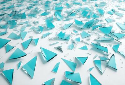 The primary subject of the image is a collection of broken glass pieces, predominantly blue in color. These pieces are scattered across a white surface, creating a visually striking and somewhat chaotic scene. The setting appears to be a workspace or an artistic environment where these broken glass pieces have been gathered and displayed. The visual style of the image is a mix of realism and artistic expression, as the broken glass pieces are not only aesthetically pleasing but also evoke a sense of fragility and impermanence.