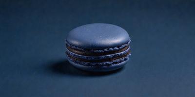 The primary subject of the image is a small, round, blue macaron with a chocolate filling. The setting is a dark blue background, which creates a contrast with the blue and brown colors of the macaron. The visual style is a close-up of the macaron, showcasing its details. The colors and mood of the image are predominantly dark and moody, with the blue macaron standing out against the dark blue background.