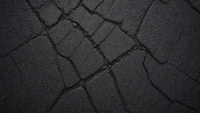 The primary subject of the image is a black, cracked, and broken pavement. The setting is a dark, possibly nighttime, environment. The visual style is a close-up of the pavement, showcasing its texture and details. The colors and mood are dark and moody, with the black pavement and the nighttime setting creating a sense of unease or mystery.