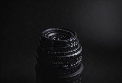 The primary subject of the image is a close-up of a camera lens. The lens is black and appears to be a prime lens. The setting is a dark room, which creates a moody atmosphere. The visual style is a realistic representation of the camera lens, and the colors are predominantly black, giving the image a dramatic and artistic feel.