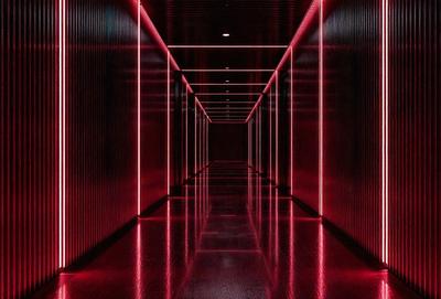 The primary subject of the image is a long, narrow hallway with red walls and a red floor. The setting is a dark room, and the visual style appears to be a photo. The dominant colors in the image are red, creating a bold and striking atmosphere.