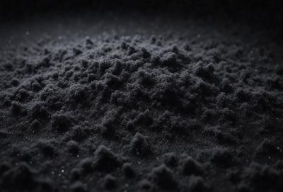 The primary subject of the image is a pile of black powder, which appears to be a dark, granular substance. The setting is a dark background, which contrasts with the black powder. The visual style of the image is a close-up of the black powder, showcasing its texture and appearance. The colors and mood of the image are predominantly dark, with the black powder being the main focus.