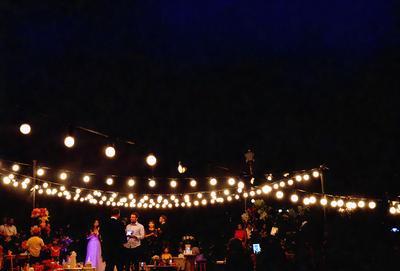 The image features a group of people gathered under a canopy, possibly at a wedding or a similar event. There are at least 13 people visible in the scene, with some standing closer to the foreground and others further back. The people are engaged in conversation and enjoying the outdoor setting.

The canopy is adorned with numerous lights, creating a warm and inviting atmosphere. The lights are scattered throughout the scene, with some closer to the people and others further away. The combination of the people, the