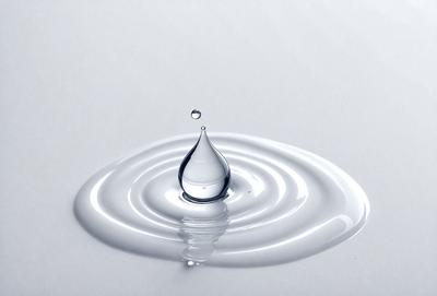 The primary subject of the image is a single drop of water, which is captured in a close-up shot. The setting is a white surface, which could be a wall or a table. The visual style is a realistic representation of the water drop, and the colors are predominantly white, giving the image a clean and minimalist appearance.