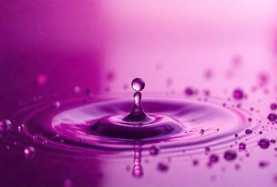 The primary subject of the image is a single drop of water, which is floating on a purple surface. The setting appears to be a dark room, and the visual style is a close-up of the drop of water. The colors and mood of the image are predominantly purple, creating a calm and serene atmosphere.