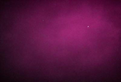 The primary subject of the image is a purple sky with a moon visible in the background. The setting is a dark, cloudy night with a pinkish hue. The visual style is a combination of a photo and digital art, creating a unique and artistic atmosphere. The colors and mood of the image are predominantly purple and dark, with a hint of pink, giving it a moody and dramatic feel.