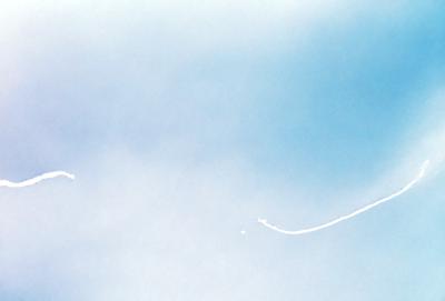 The primary subject of the image is a rocket flying through the sky. The setting is a cloudy blue sky, and the visual style appears to be a photo. The colors in the image are predominantly blue and white, creating a calm and serene atmosphere.