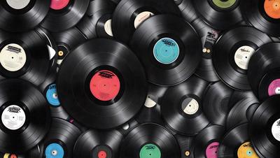 The primary subject of the image is a collection of various colored vinyl records. The records are arranged in a visually appealing manner, creating a patterned background. The setting is a room filled with these records, showcasing the different colors and designs of the records. The visual style of the image is a close-up of the records, emphasizing their unique colors and patterns. The overall atmosphere of the image is vibrant and lively, reflecting the diverse and creative nature of the vinyl records.