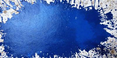 The primary subject of the image is a blue splash of paint, which is the main focus of the scene. The setting is a white surface, which serves as the background. The visual style of the image is a close-up of the blue splash, creating a sense of depth and detail. The dominant colors in the image are blue and white, giving it a calm and serene atmosphere.