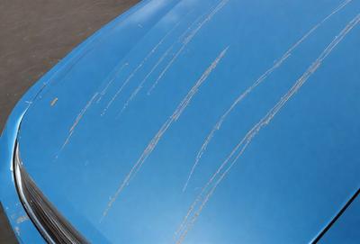 The primary subject of the image is a blue car with a scratched surface. The setting is outdoors, and the visual style appears to be a photo. The car has a blue hue, and the scratches on the surface create a mood of damage or wear.