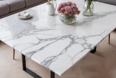 The primary subject of the image is a white marble table with a vase of flowers on top. The table is placed in a setting with a couch in the background. The visual style of the image is a photo, and the colors and mood are predominantly white, giving the scene a clean and elegant appearance.