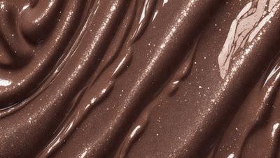The primary subject of the image is a close-up of a chocolate-colored surface with a pattern of swirls and waves. The setting appears to be a dark room, and the visual style is a close-up of the chocolate surface. The colors and mood of the image are predominantly dark and moody, with the chocolate color adding a rich and luxurious feel to the scene.