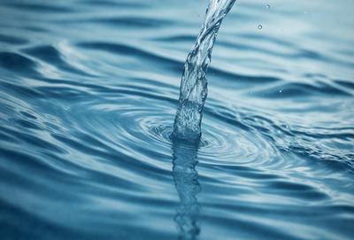 The primary subject of the image is a water droplet, which is captured in mid-air as it falls into the water. The setting is a body of water, possibly an ocean, with a clear blue color. The visual style is a close-up of the water droplet, showcasing its details and texture. The colors and mood of the image are serene and calming, as the water droplet creates a peaceful and tranquil atmosphere.