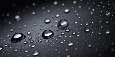 The primary subject of the image is a close-up of water droplets on a black surface. The setting is a dark background, which contrasts with the bright droplets. The visual style is a black and white photo, which adds a timeless and classic feel to the image. The colors and mood are predominantly dark, with the water droplets being the main source of light and interest in the scene.