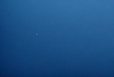 The primary subject of the image is a single moon, which is visible in the sky. The setting is a vast, dark blue sky, with no clouds or other objects visible. The visual style is a realistic representation of the moon and the sky. The colors and mood of the image are predominantly dark and moody, with the blue sky providing a contrasting backdrop to the luminous moon.
