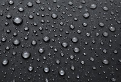 The primary subject of the image is a close-up of a black surface with numerous small droplets of water on it. The setting is a dark background, and the visual style appears to be a photo. The colors and mood of the image are predominantly dark, with the droplets of water being the main focus.