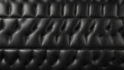 The primary subject of the image is a black leather jacket with a patterned design. The setting is a black background, which highlights the jacket's texture and pattern. The visual style is a close-up of the jacket, showcasing its details. The colors and mood of the image are dark and moody, emphasizing the leather jacket's sophisticated and stylish appearance.