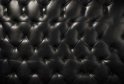 The primary subject of the image is a black leather couch. The setting is a black background, which emphasizes the couch's design. The visual style is a close-up of the couch, showcasing its texture and details. The colors and mood of the image are predominantly dark, creating a sense of luxury and sophistication.