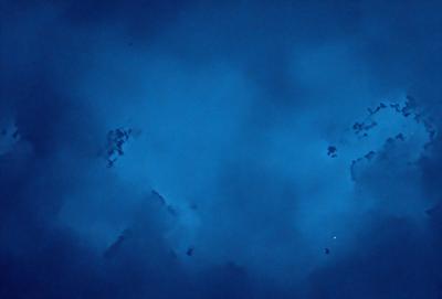 The primary subject of the image is a cloudy blue sky. The setting is a vast expanse of the sky, with no visible land or other objects. The visual style is a natural, outdoor scene. The colors and mood are predominantly blue, with a slightly darker tone, creating a serene and peaceful atmosphere.