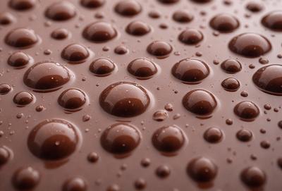 The primary subject of the image is a close-up of a chocolate surface with water droplets on it. The setting is a dark background, which contrasts with the chocolate and water droplets. The visual style is a close-up, which emphasizes the details of the chocolate and water droplets. The colors are predominantly brown and black, with the chocolate being the dominant color, and the water droplets adding a contrasting element. The overall atmosphere is moist and