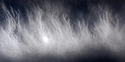 The primary subject of the image is a cloudy sky with a sun shining through it. The setting is a cloudy day, and the visual style is a photo. The colors and mood of the image are predominantly dark and moody, with the sunlight creating a contrasting effect.