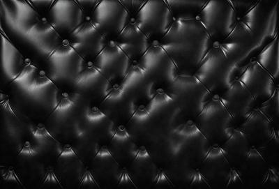 The primary subject of the image is a black leather couch. The setting is a black background, which emphasizes the couch's color and texture. The visual style is a close-up of the couch, showcasing its details. The colors and mood of the image are dark and moody, with the black leather couch being the main focal point.