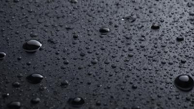 The primary subject of the image is a close-up of a black surface with numerous droplets of water on it. The setting is a dark background, and the visual style appears to be a photo. The colors in the image are predominantly black, with the droplets of water being the main focus and creating a moist and wet atmosphere.