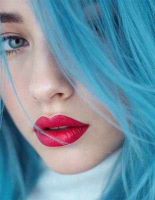 The primary subject of the image is a woman with blue hair and red lipstick. She is wearing a white shirt and has her hair blowing in the wind. The setting is outdoors, with a natural background. The visual style is a close-up portrait, capturing the woman's features and expression. The colors and mood of the image are vibrant and energetic, with the blue hair and red lipstick creating a striking contrast against the natural backdrop.