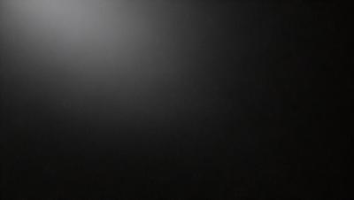 The primary subject of the image is a black background with a white light source. The setting is a dark room, and the visual style is a black and white photo. The colors and mood of the image are predominantly dark and moody, with the white light source creating a stark contrast against the black background.