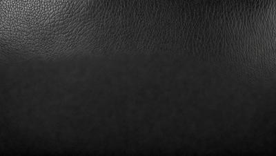 The primary subject of the image is a black leather jacket. The setting is a black background, which creates a contrast with the leather jacket. The visual style is a close-up of the leather jacket, showcasing its texture and details. The colors and mood of the image are dark and moody, emphasizing the leather jacket's sleek and stylish appearance.