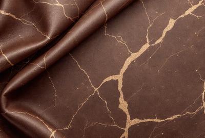 The primary subject of the image is a piece of brown leather with a pattern of gold and brown spots. The leather is laid out on a table, and it appears to be a close-up of the material. The setting is indoors, with the leather as the main focus of the scene. The visual style is a close-up of the leather, showcasing its texture and pattern. The colors and mood of the image are warm and earthy, with the gold and brown spots adding a touch