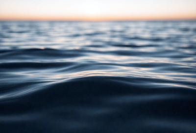The primary subject of the image is the ocean, with its vast expanse of water and the sun setting in the background. The setting is a serene beach, and the visual style is a photo of the ocean. The colors and mood of the image are predominantly blue, with a hint of orange from the sunset, creating a peaceful and calming atmosphere.