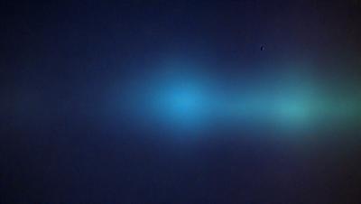 The primary subject of the image is a blue light, which appears to be a neon light or a light source in the distance. The setting is a dark, foggy sky, creating a moody atmosphere. The visual style of the image is a photo, capturing the blue light against the dark background. The colors and mood of the image are predominantly dark and mysterious, with the blue light providing a contrasting element.