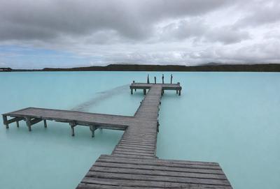 The primary subject of the image is a pier extending out into the water. The pier is made of wood and has a docking area. The setting is a large body of water, possibly an ocean, with a cloudy sky above. The visual style is a photo, capturing the pier and its surroundings in a natural environment. The colors and mood of the image are predominantly blue, with a calm and serene atmosphere.